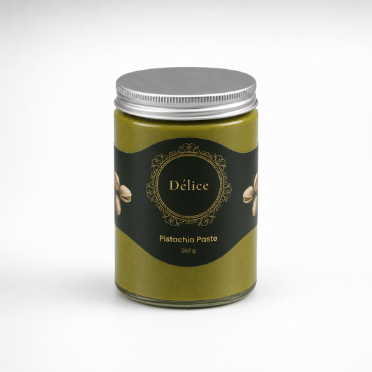 A glass jar of Pistachio Paste by Délice, featuring a dark olive green paste inside, a black label with gold details, and a silver metal lid, set against a clean white background.