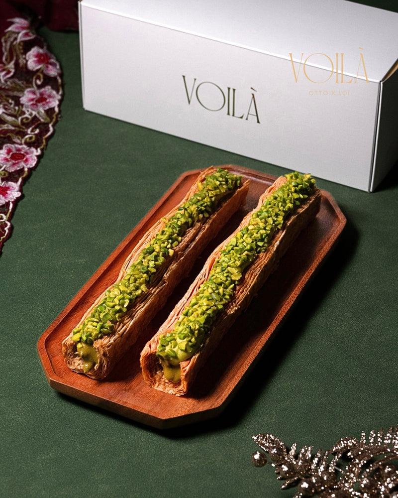 Two pastries with green pistachio granules filling on a wooden tray in front of a VOILA box.